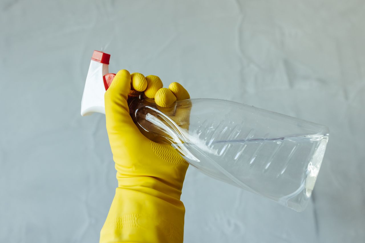 Services Close-up of a hand wearing yellow gloves holding a transparent spray bottle, isolated on a plain background.