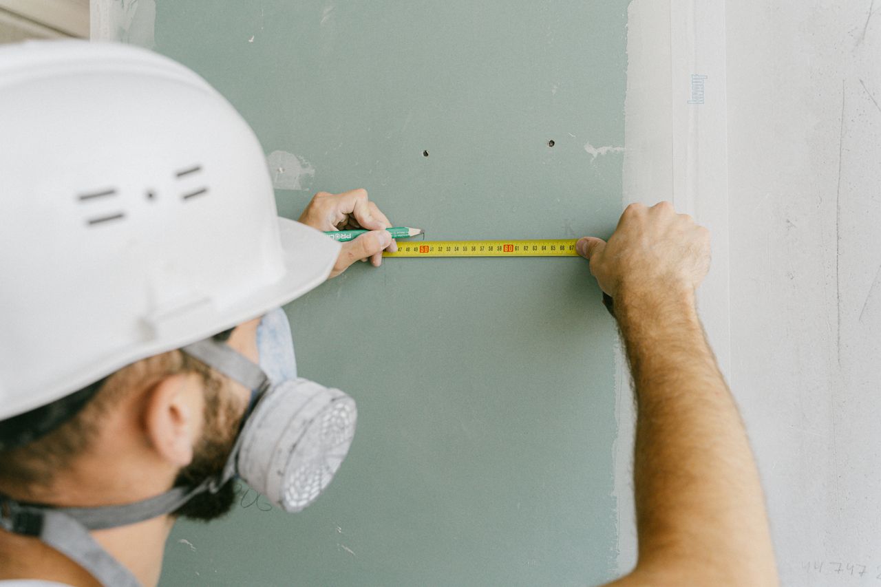 Services A construction worker precisely measuring drywall, wearing a helmet and mask for safety.